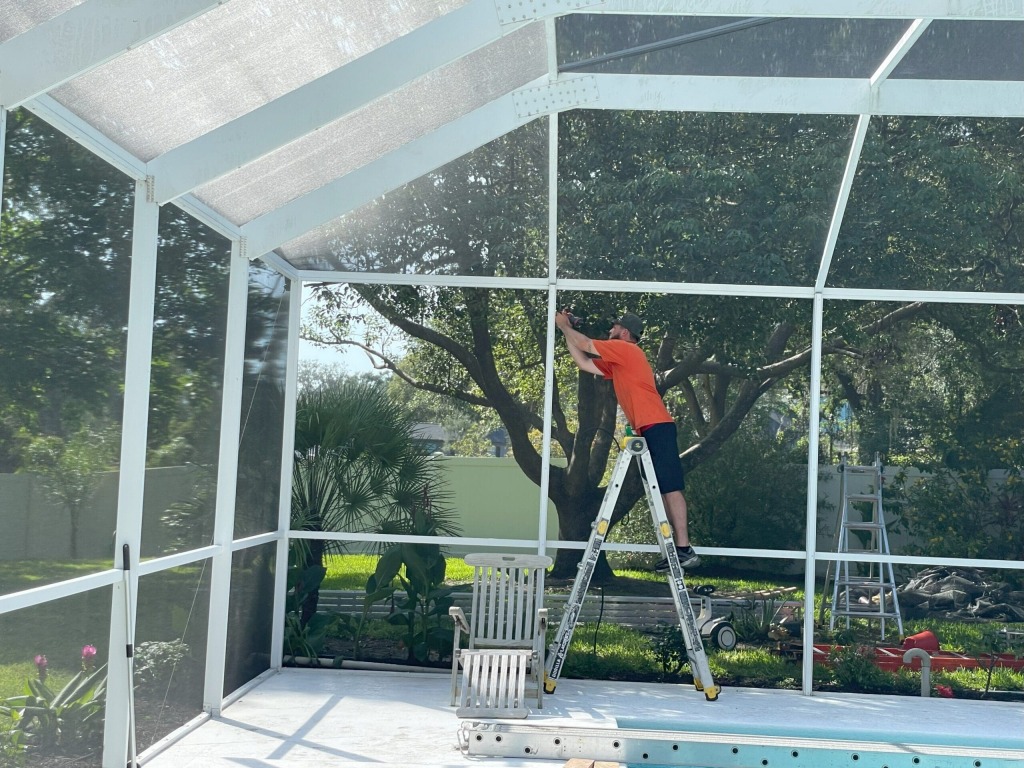 Man repairing pool enclosure screen on ladder in backyard patio area