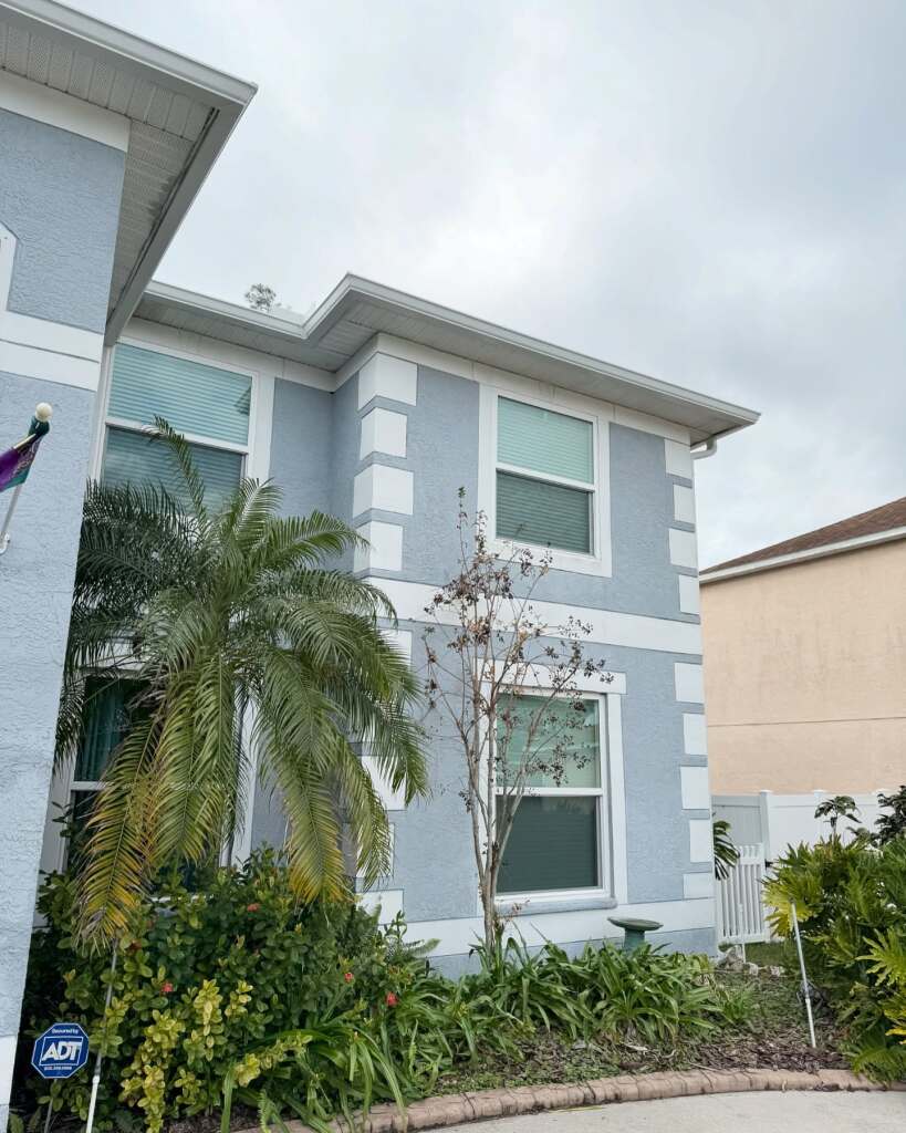 Two-story light blue house with lush greenery and palm tree