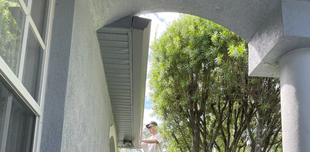 Man cleaning house gutters under a sunny archway with green trees nearby.