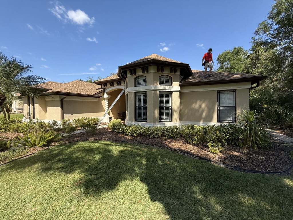 Man in red shirt inspecting roof of beige house on a clear sunny day